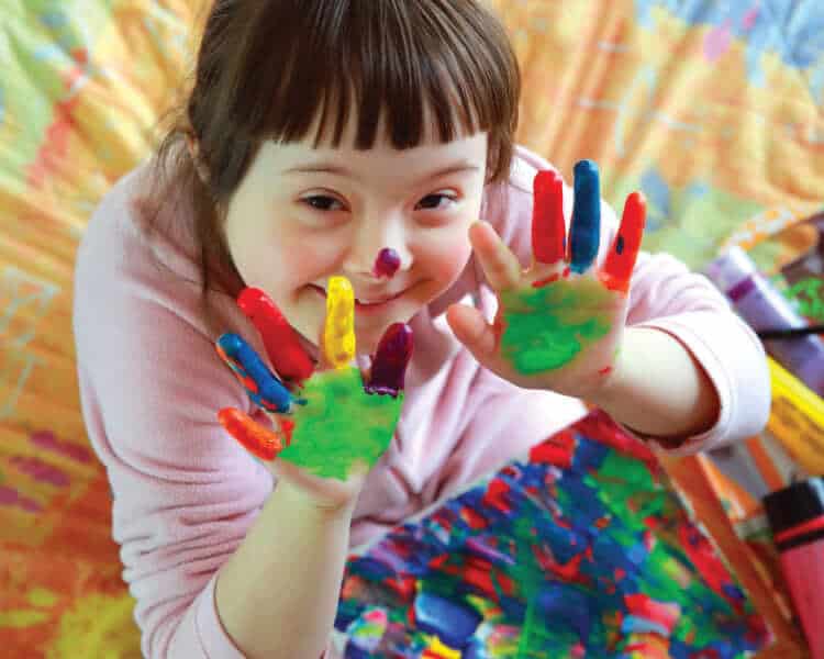 一个有特殊需要的孩子从地板上抬起头，手上沾着颜料。她在学校微笑着画手指画。