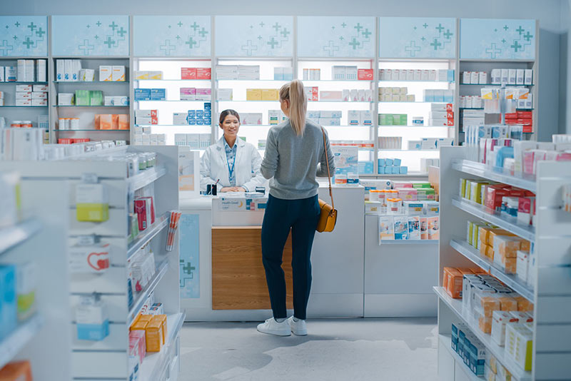 药店过道的广角镜头，尽头是药房柜台。药剂师正在帮一位妇女拿优惠券和处方。