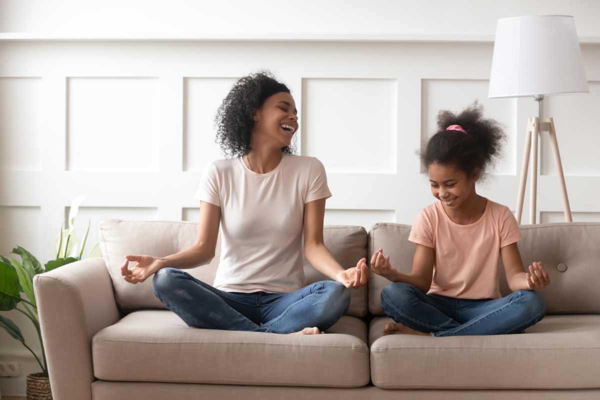Mixed-race mother little daughter distracted from meditation laughing sit in lotus position on couch in living room, mom teach kid beginner yoga asana, healthy life habits, activities at home