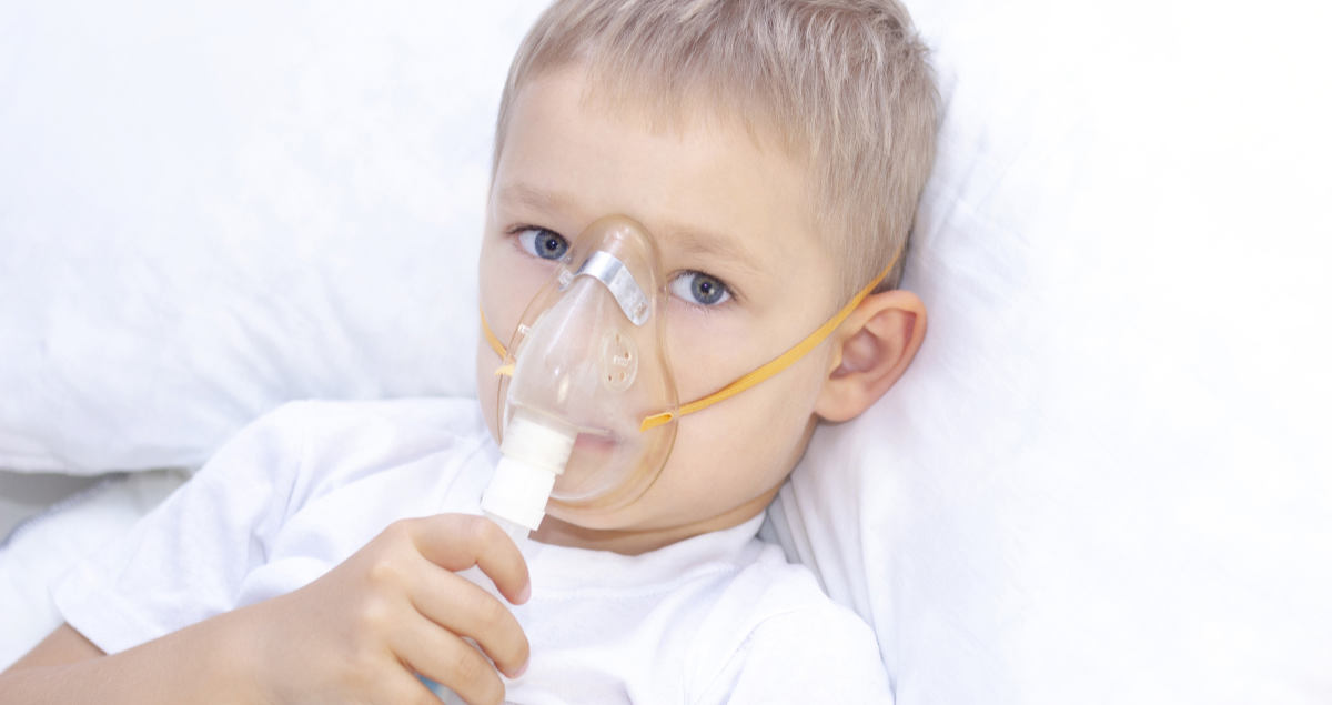 Photo of boy laying in bed holding a mask for a respiratory treatment due to a nighttime asthma attack.