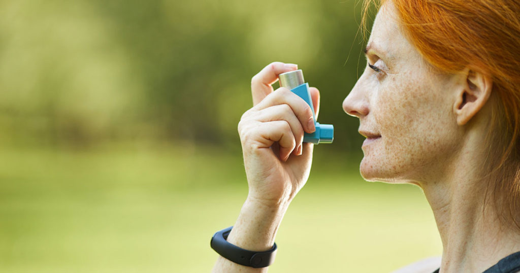 Woman with red hair holding an asthma inhaler close to her mouth like she is ready to use it. There is a blurry image of green grass and trees in the background indicating summer.