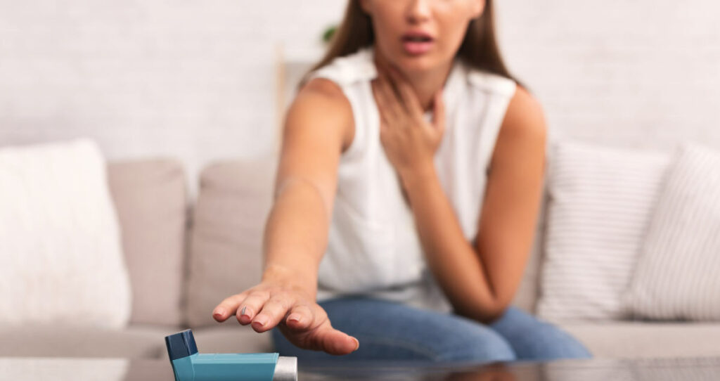 Middle aged woman sitting on her couch and reaching for her asthma inhaler with one hand while holding her airways with the other hand.