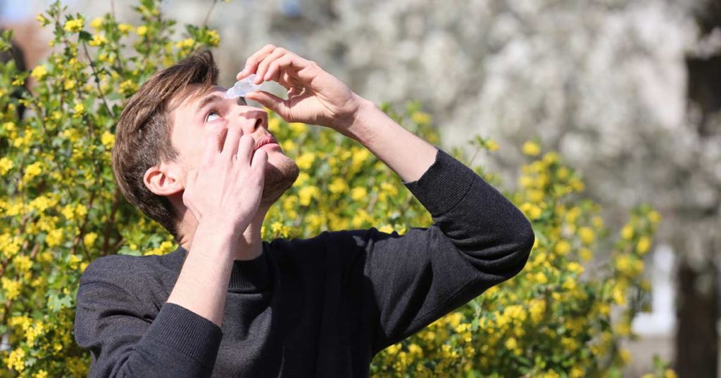 Adult man putting eye drops in his eyes, with trees in the background causing his discomfort.