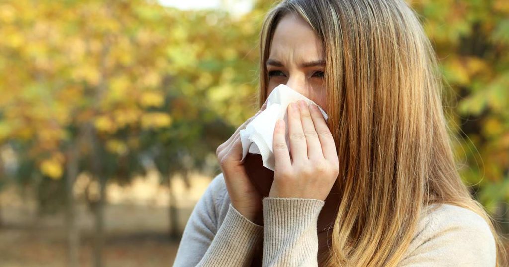 Adult woman sneezing into a tissue. She is in a park with trees producing pollen in the background.