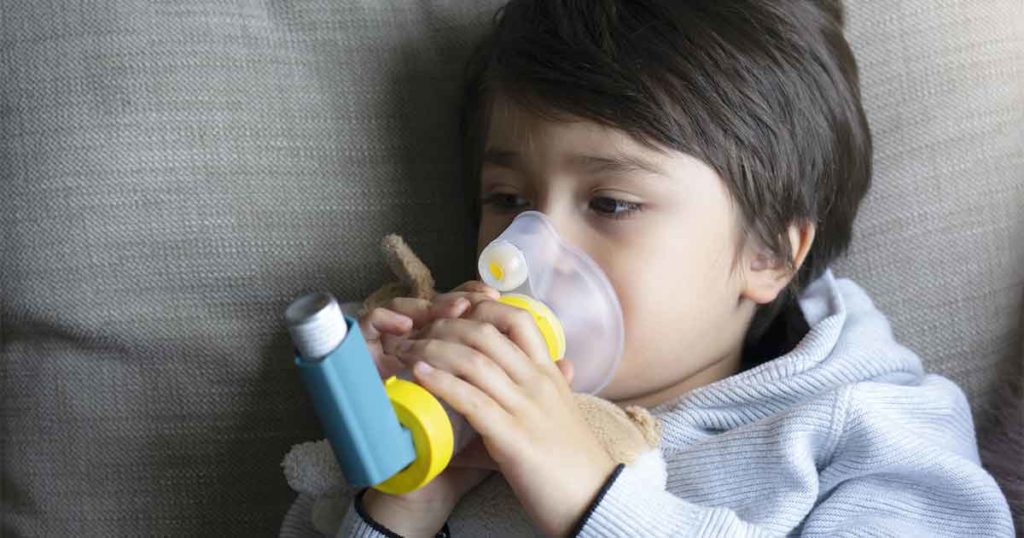 Young boy laying down using an asthma inhaler with a spacer.