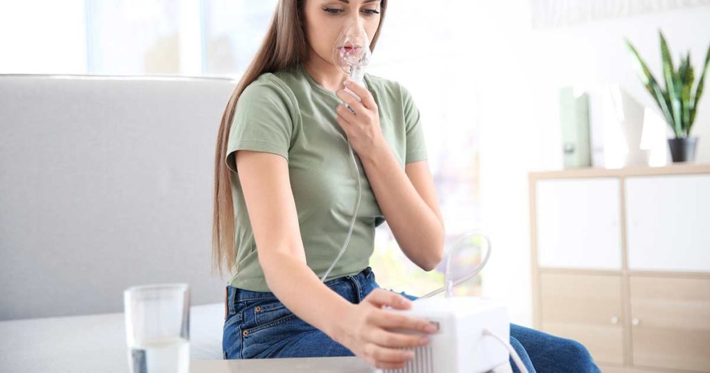 Woman using a nebulizer in her living room. She is holding her chest during her asthma treatment.